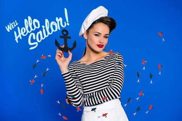 Portrait of fashionable young woman in retro clothing with anchor in hand and well hello sailor inscription isolated on blue — Stock Photo
