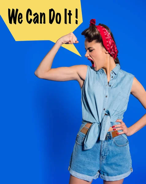 Young woman in retro clothing showing muscles and shouting with we can do it speech bubble isolated on blue — Stock Photo