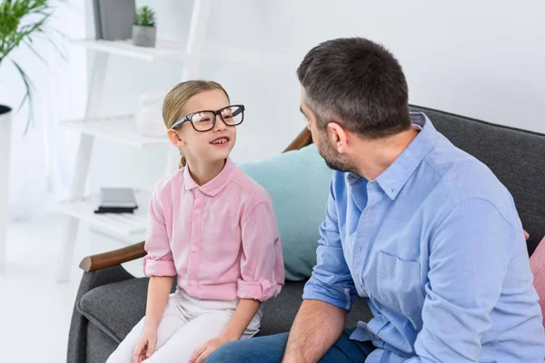 Father and daughter in eyeglasses looking at each other while sitting on sofa at home — Stock Photo