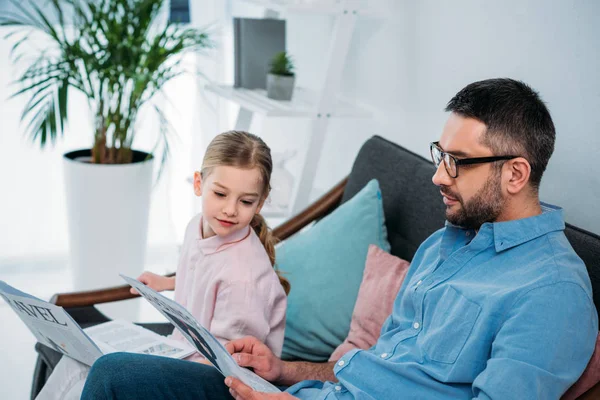 Vista lateral del padre y la hija con periódicos descansando en el sofá en casa — Stock Photo