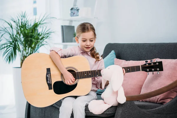 Retrato de garoto bonito tocando guitarra acústica enquanto sentado no sofá em casa — Fotografia de Stock