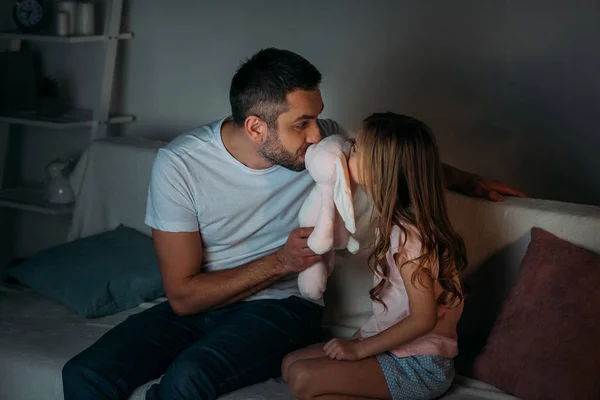 Батько і маленька дочка цілують іграшку, сидячи на дивані вдома — стокове фото