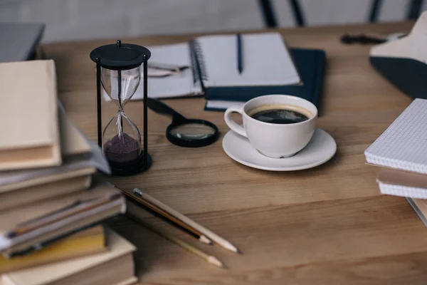 Крупный план чашки кофе и песочных часов на рабочем столе с письменными принадлежностями — стоковое фото