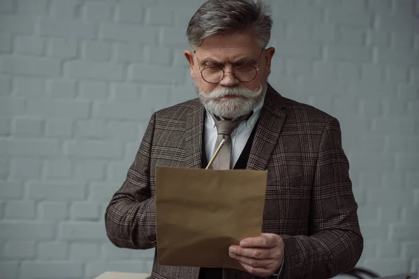 Thoughtful senior man with pencil and sheet of paper in front of white brick wall — Stock Photo