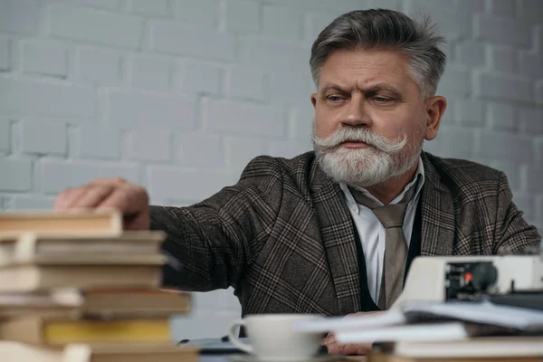 Close-up shot of bearded senior writer at workplace taking book from stack — Stock Photo