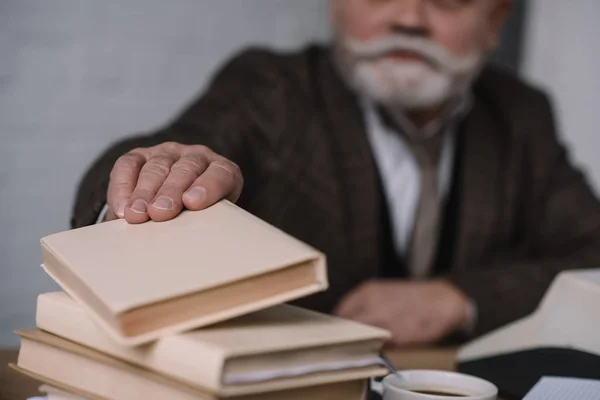 Close-up shot of senior writer at workplace taking book from stack — Stock Photo