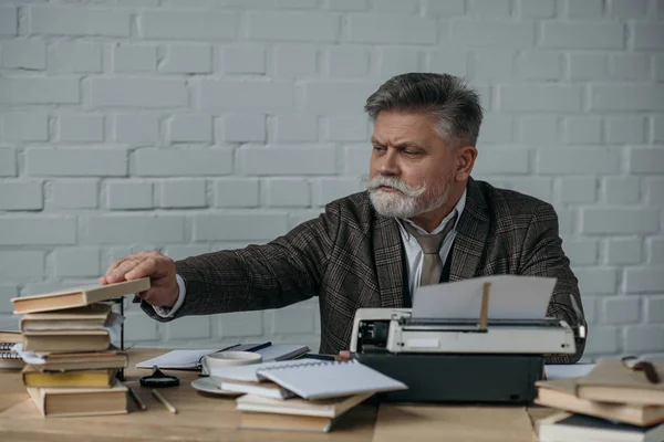 Senior writer at workplace taking book from stack — Stock Photo