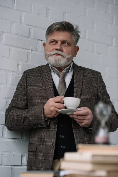 Reflexivo hombre mayor bebiendo café con pila de libros y reloj de arena en primer plano — Stock Photo