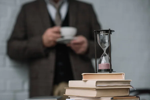 Close-up shot of stack of books and hourglass with man drinking coffee blurred on background — Stock Photo