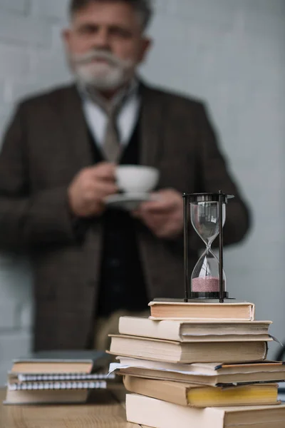 Blurred senior man drinking coffee with stack of books and hourglass on foreground — Stock Photo