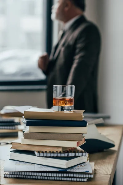Close-up shot of glass of whiskey on stack of books with senior man standing blurred on background — Stock Photo
