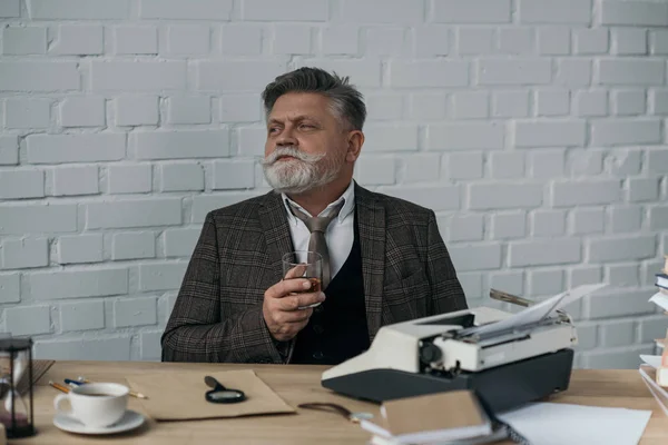 Felice scrittore anziano con un bicchiere di whisky sul posto di lavoro — Foto stock