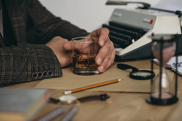 Recortado tiro de escritor senior con vaso de whisky en el lugar de trabajo - foto de stock