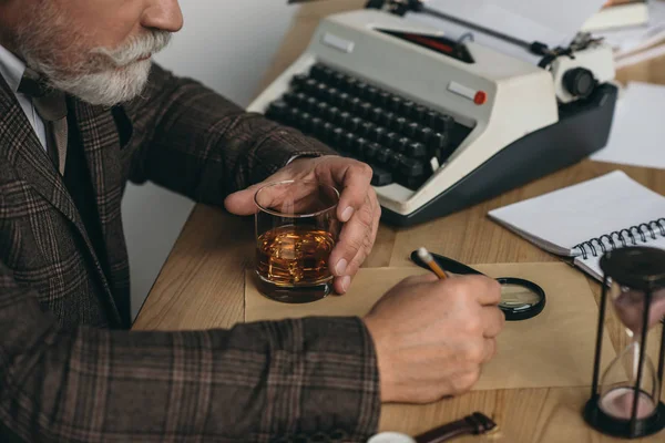 Recortado tiro de escritor senior trabajando en manuscrito y sosteniendo vaso de whisky - foto de stock
