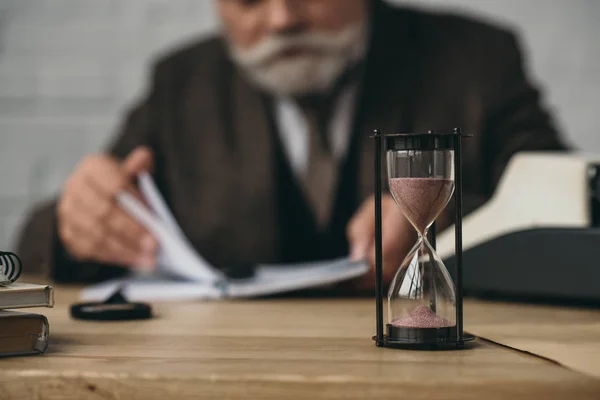 Senior writer working with hourglass standing on foreground — Stock Photo