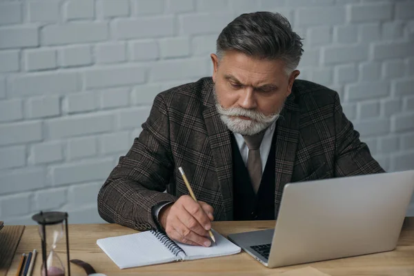 Escritor sênior bonito trabalhando com laptop e fazer anotações no notebook — Fotografia de Stock