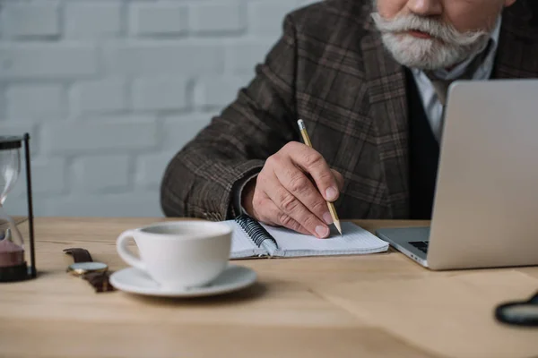 Recortado tiro de escritor senior que trabaja con el ordenador portátil y toma notas en el cuaderno - foto de stock