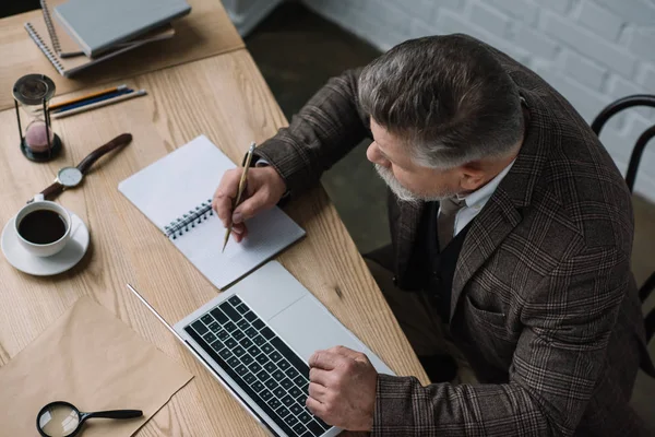 High angle view of senior writer working with laptop and making notes in notebook — Stock Photo