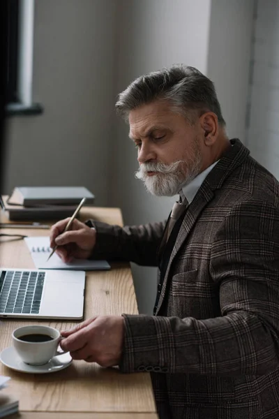 Scrittore anziano serio che lavora con computer portatile e prende appunti in notebook — Foto stock