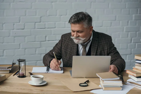 Felice scrittore anziano che lavora con il computer portatile e prendere appunti nel notebook — Foto stock