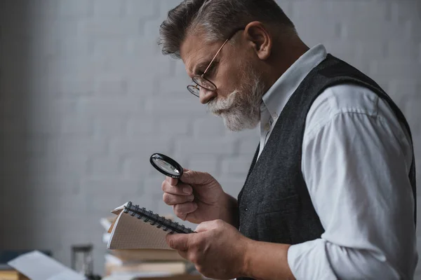 Handsome senior man reading notes in notebook with magnifying glass — Stock Photo