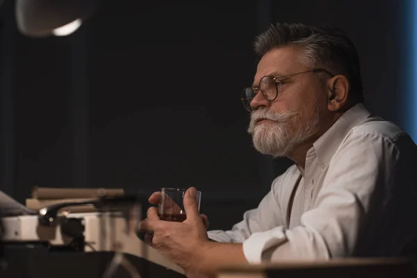 Pensativo escritor senior sentado en el lugar de trabajo con un vaso de whisky aislado en negro - foto de stock