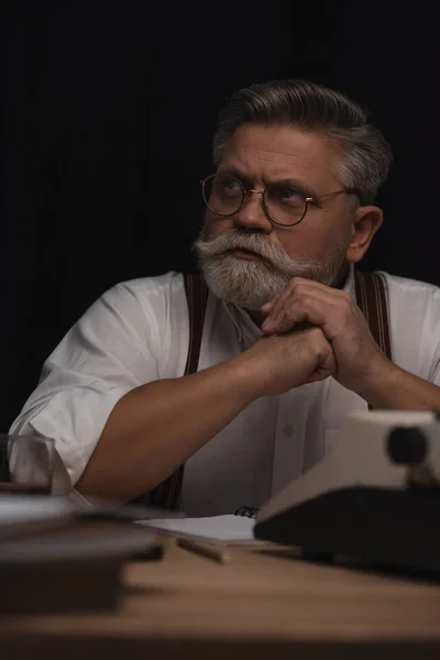 Thoughtful senior writer sitting at workplace and looking away isolated on black — Stock Photo