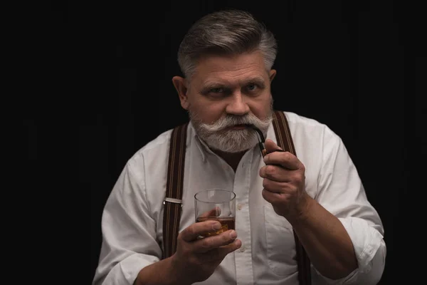 Mature homme fumeur pipe et boire whisky isolé sur noir — Photo de stock