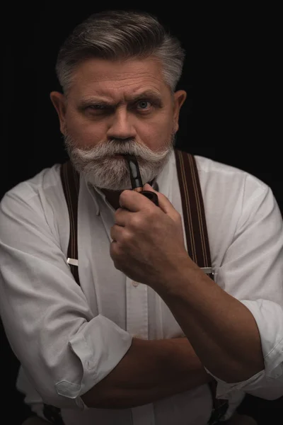 Mature man smoking pipe and looking at camera isolated on black — Stock Photo