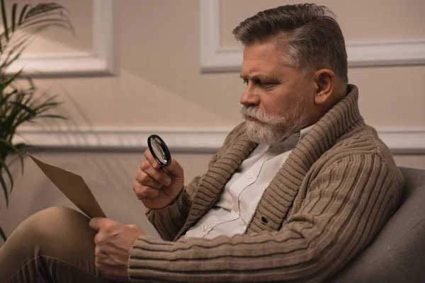 Senior man reading letter with magnifying glass while sitting in armchair — Stock Photo