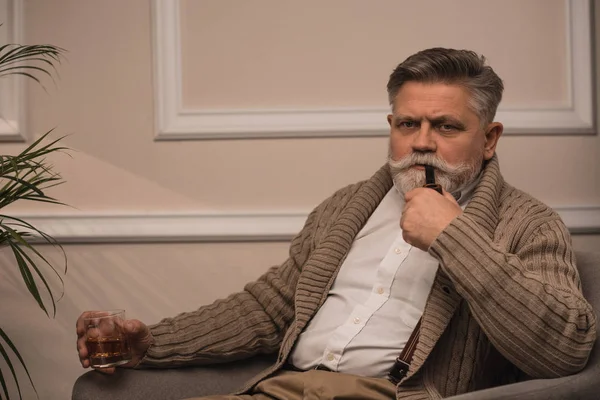 Handsome senior man smoking pipe in armchair and looking at camera — Stock Photo