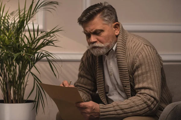 Thoughtful senior man in sweater writing letter while sitting in armchair — Stock Photo