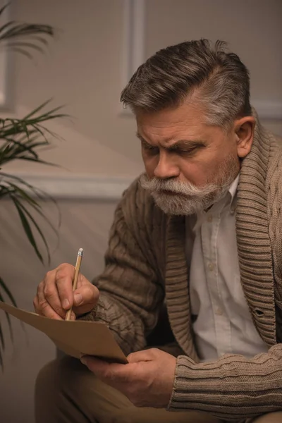 Вдумчивый пожилой мужчина в свитере пишет письмо — стоковое фото