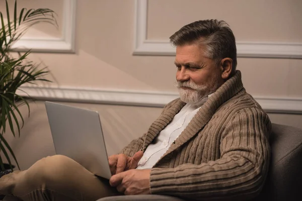 Hombre mayor feliz usando el ordenador portátil mientras está sentado en el sillón - foto de stock