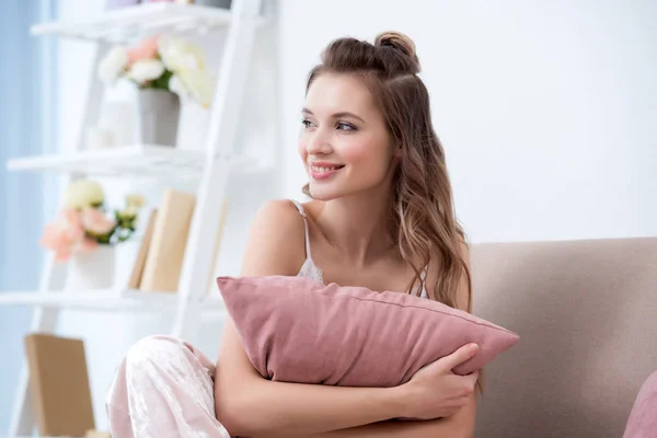 Bella ragazza sorridente in pigiama tenendo cuscino e guardando lontano a casa — Foto stock