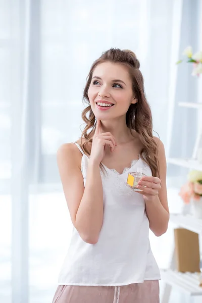 Красива усміхнена молода жінка в піжамі тримає пляшку парфумів і дивиться — стокове фото