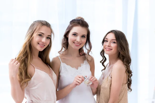 Beautiful young women in pajamas holding bottle of perfume and smiling at camera — Stock Photo