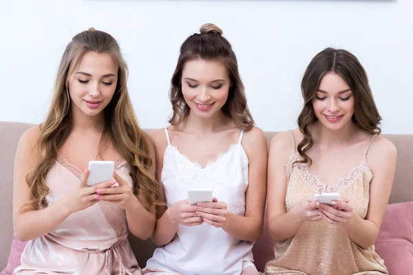 Hermosas mujeres jóvenes sonrientes en pijama sentadas juntas y usando teléfonos inteligentes - foto de stock