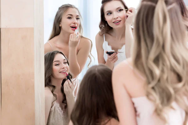 Beautiful happy young women in pajamas applying makeup and looking at mirror — Stock Photo