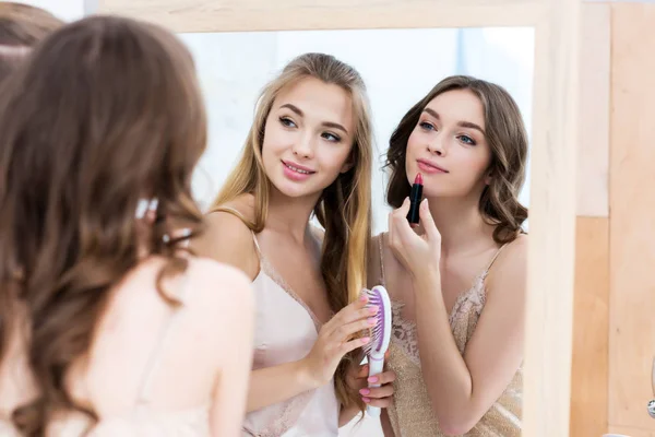 Hermosas mujeres jóvenes en pijama aplicando maquillaje y mirando al espejo - foto de stock