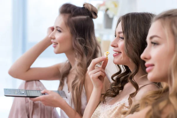 Side view of beautiful smiling girlfriends in pajamas eating popcorn and watching tv together — Stock Photo
