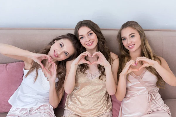 Hermosas mujeres jóvenes en pijama mostrando símbolo del corazón de la mano y sonriendo a la cámara - foto de stock