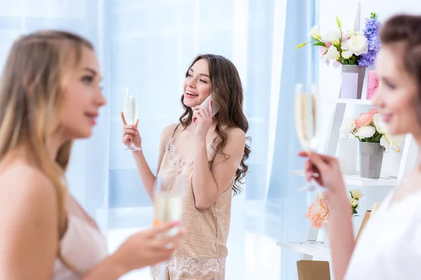 Beautiful girlfriends in pajamas drinking champagne while friend talking by smartphone — Stock Photo