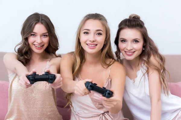 Belles jeunes femmes en pyjama jouant avec des joysticks et souriant à la caméra — Photo de stock