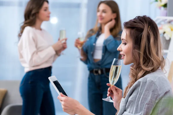 Молода жінка тримає келих шампанського і використовує смартфон, поки друзі розмовляють позаду — стокове фото