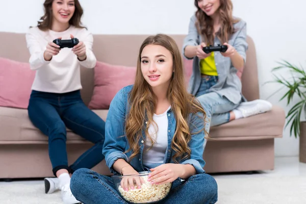 Jovem mulher comendo pipoca de tigela de vidro e olhando para a câmera enquanto amigos brincando com joysticks atrás — Fotografia de Stock