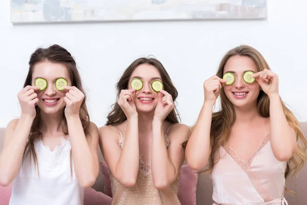 Beautiful smiling young women in pajamas holding cucumber slices near eyes — Stock Photo