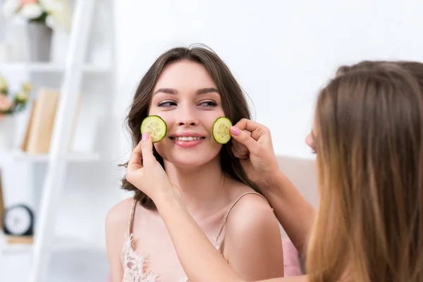 Cropped shot of girl holding slices of cucumber near face of smiling girlfriend — Stock Photo