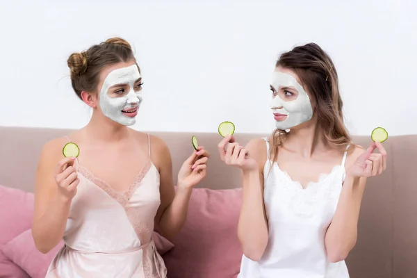 Meninas bonitas em pijama e máscaras faciais segurando fatias de pepino e olhando um para o outro — Fotografia de Stock