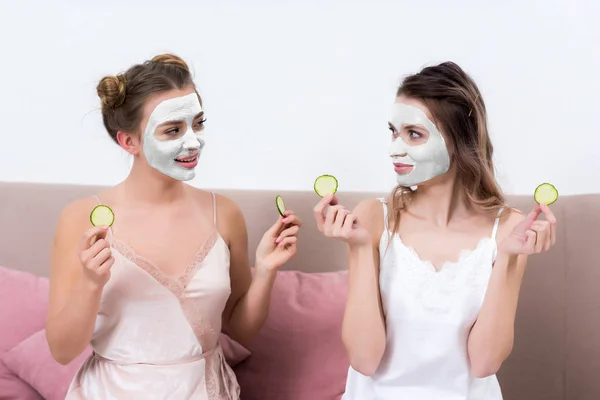 Beautiful girls in pajamas and facial masks holding slices of cucumber and looking at each other — Stock Photo